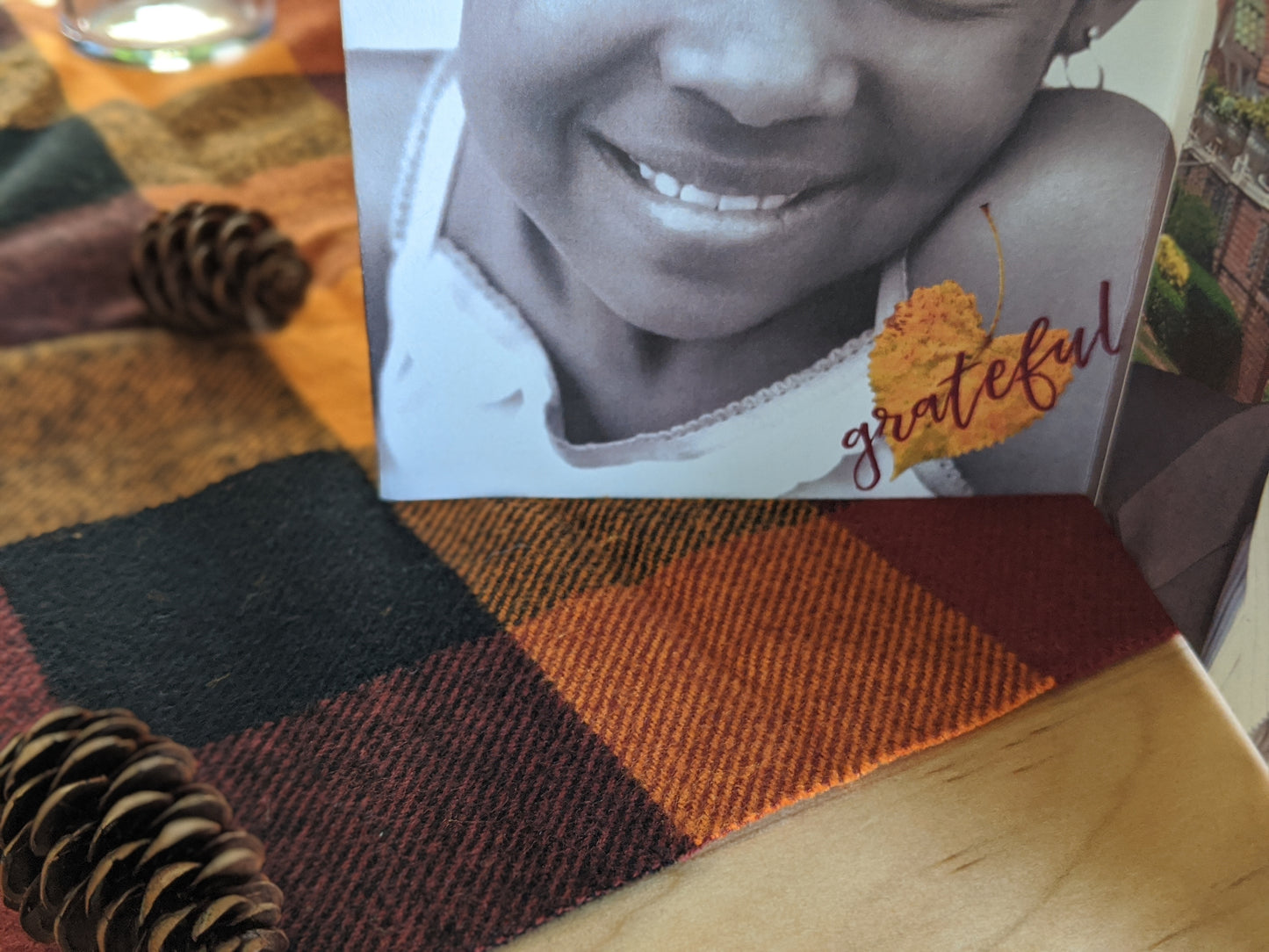 Zoomed in detail of a centerpiece sitting on a wooden table with a checkered napkin in fall colors and pinecones. There is a photo of a girl smiling collaged with a heart shaped leaf with the word grateful.