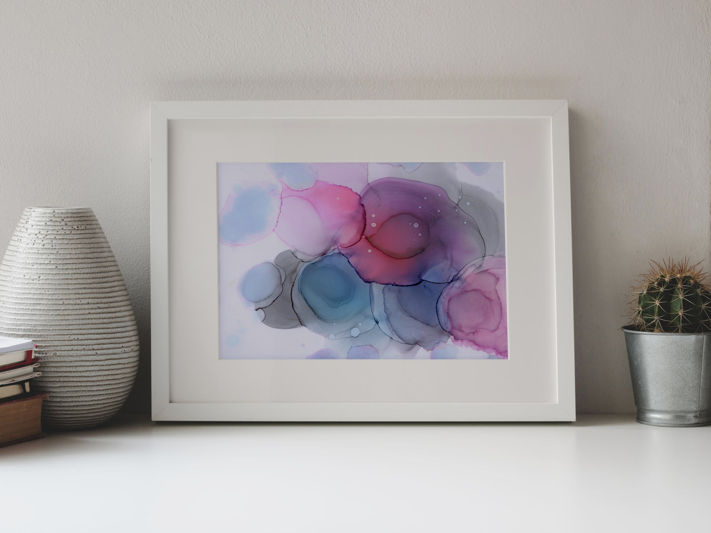 Digital mockup of Between the Stars framed in white on a white table top, leaning up against a white background, surrounded by a vase, stack of books, and tiny cactus in a pail.