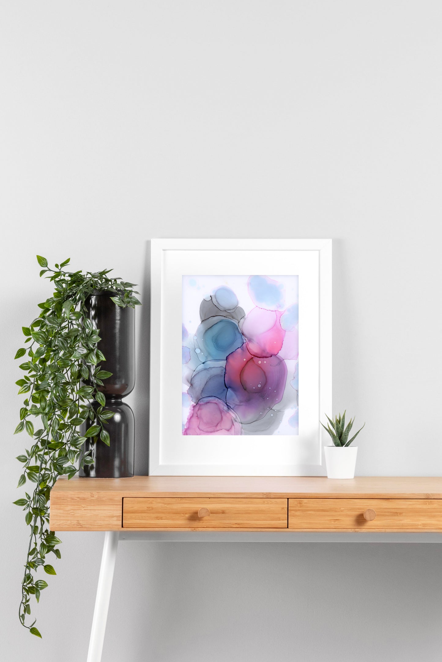 Digital mockup of Between the Stars framed in white on a wooden desk, leaning up against a neutral wall with a small succulent in a white planter on one side and greenery in a taller black planter on the other.