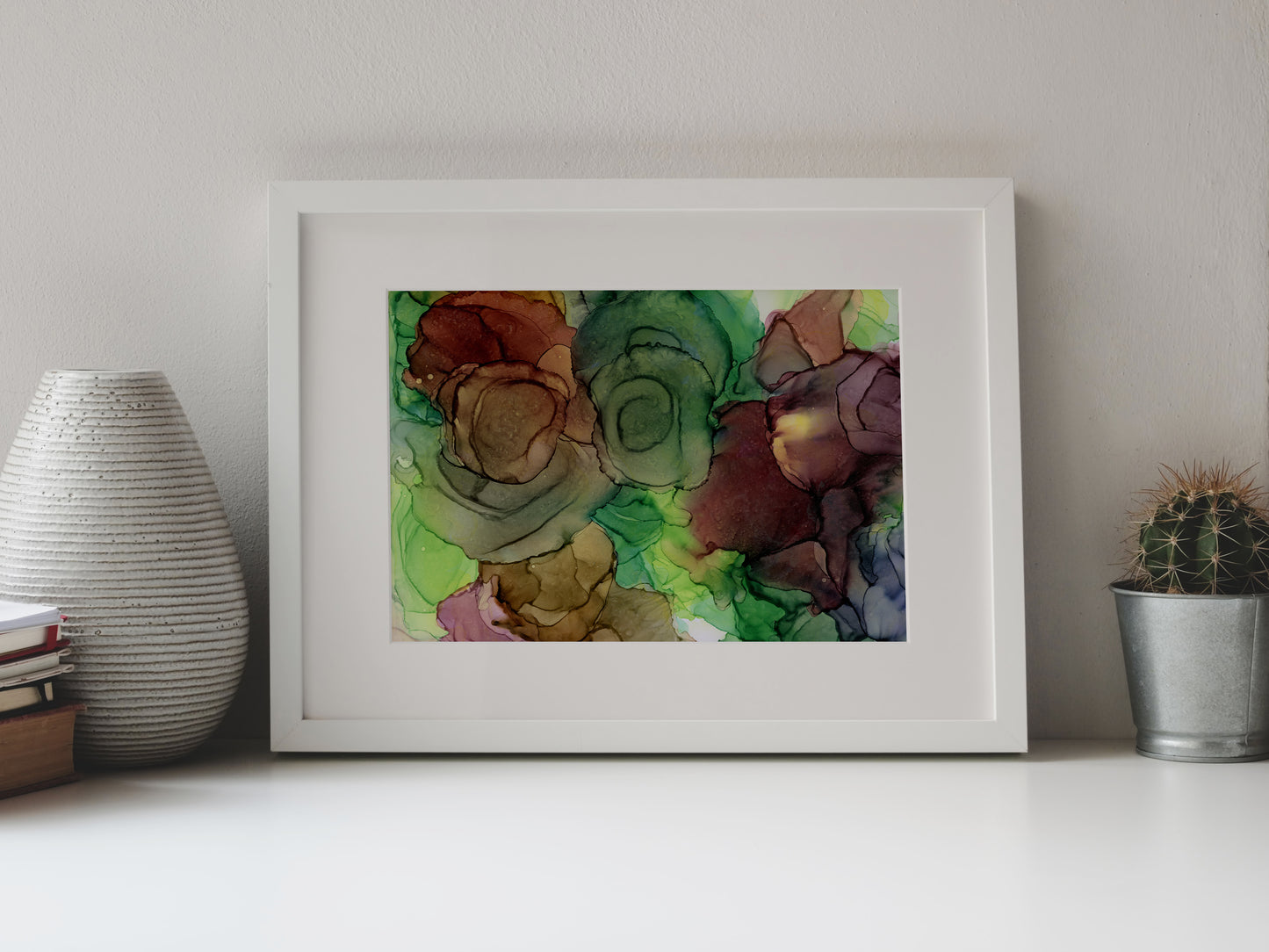 Digital mockup of Rejuvenating Blooms framed in white on a white table top, leaning up against a white background, surrounded by a vase, stack of books, and tiny cactus in a pail.