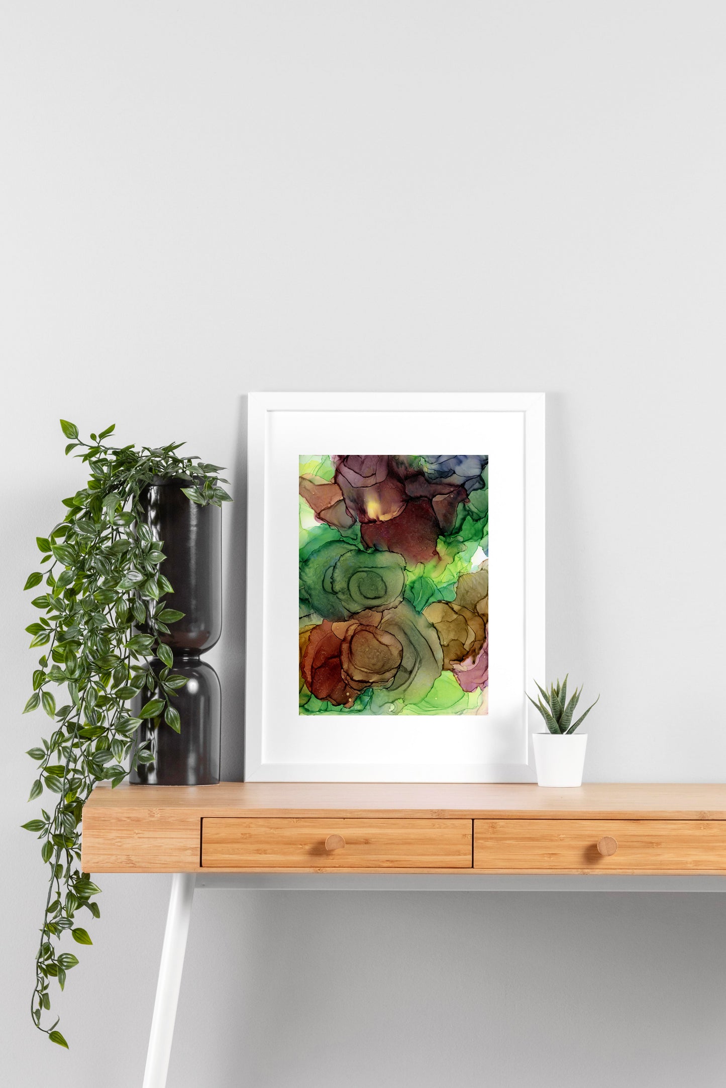 Digital mockup of Rejuvenating Blooms framed in white on a wooden desk, leaning up against a neutral wall with a small succulent in a white planter on one side and greenery in a taller black planter on the other.