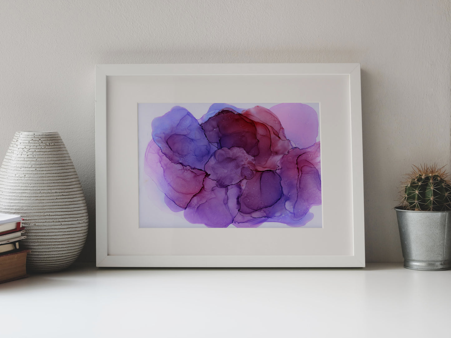Digital mockup of Bubbling Plum framed in white on a white table top, leaning up against a white background, surrounded by a vase, stack of books, and tiny cactus in a pail.
