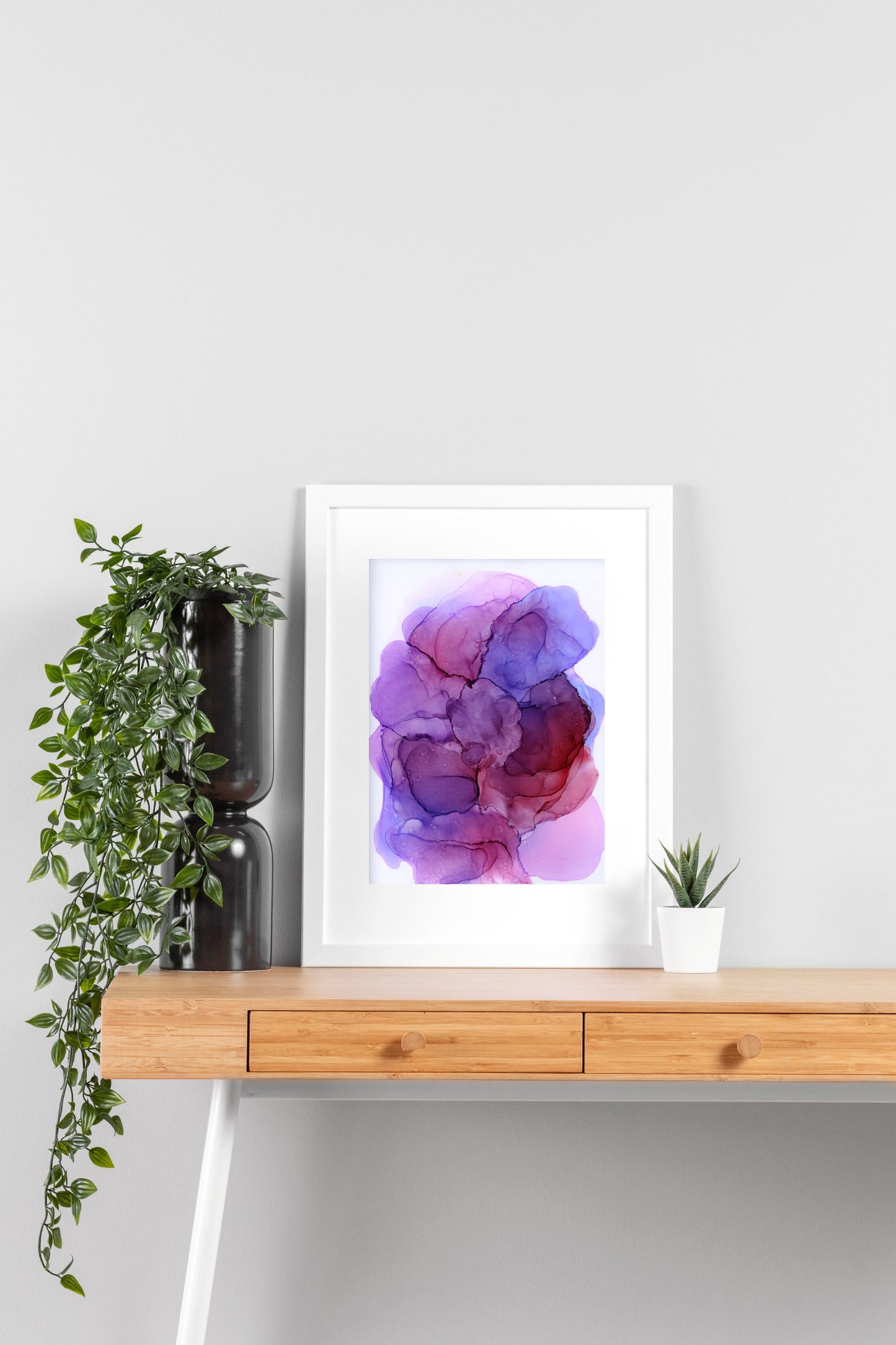 Digital mockup of Bubbling Plum framed in white on a wooden desk, leaning up against a neutral wall with a small succulent in a white planter on one side and greenery in a taller black planter on the other.