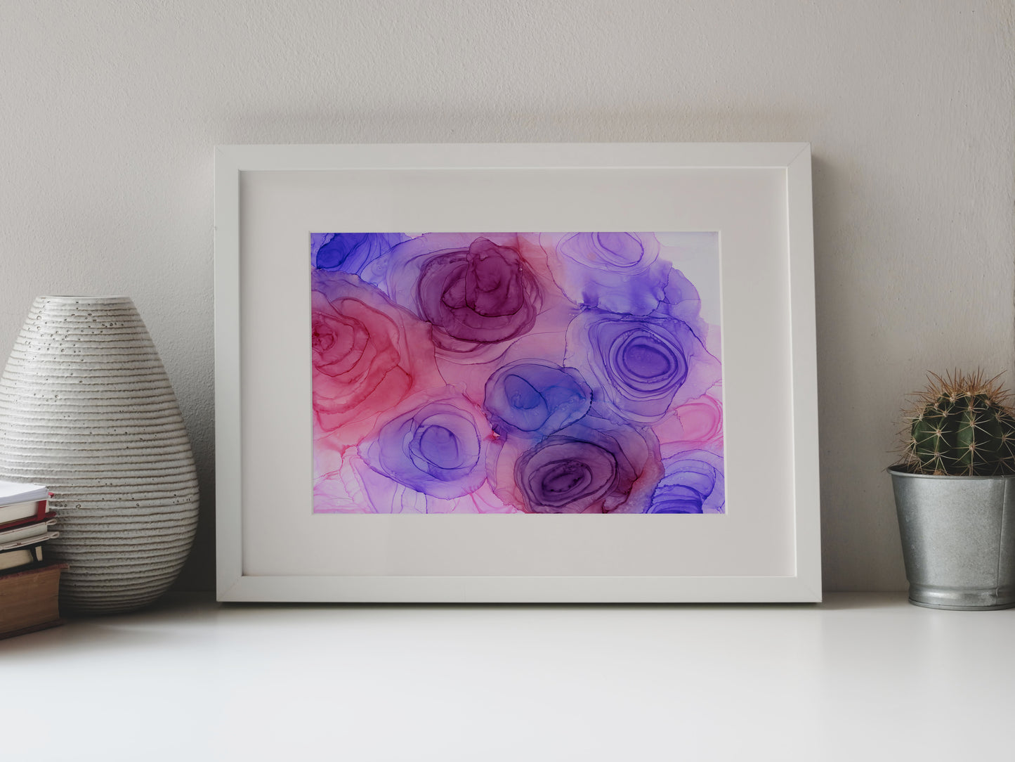 Digital mockup of Blushing Blooms framed in white on a white table top, leaning up against a white background, surrounded by a vase, stack of books, and tiny cactus in a pail.