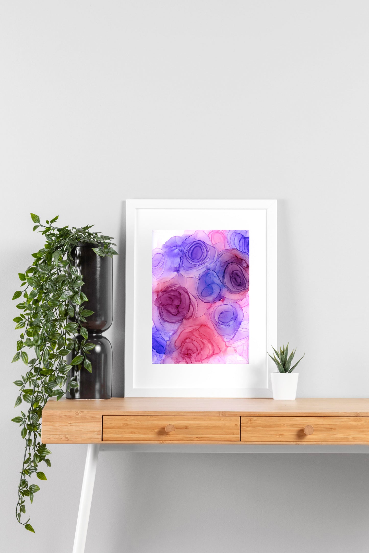 Digital mockup of Blushing Blooms framed in white on a wooden desk, leaning up against a neutral wall with a small succulent in a white planter on one side and greenery in a taller black planter on the other.