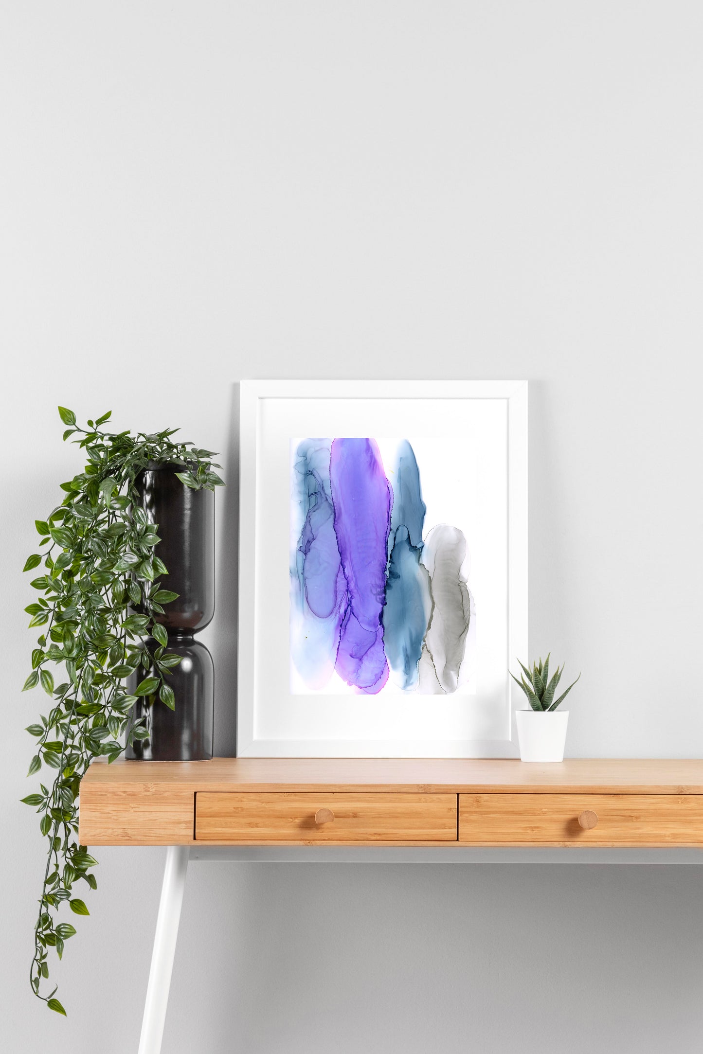Digital mockup of Layered Transitions framed in white on a wooden desk, leaning up against a neutral wall with a small succulent in a white planter on one side and greenery in a taller black planter on the other.