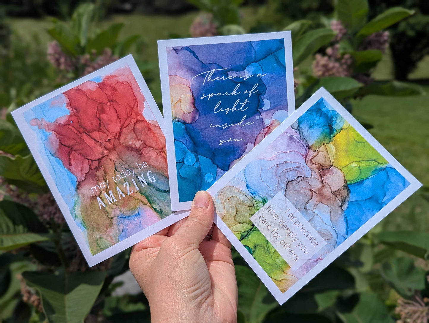 Hand holding three colorful cards with abstract ink painting designs and motivational quotes against a natural background.