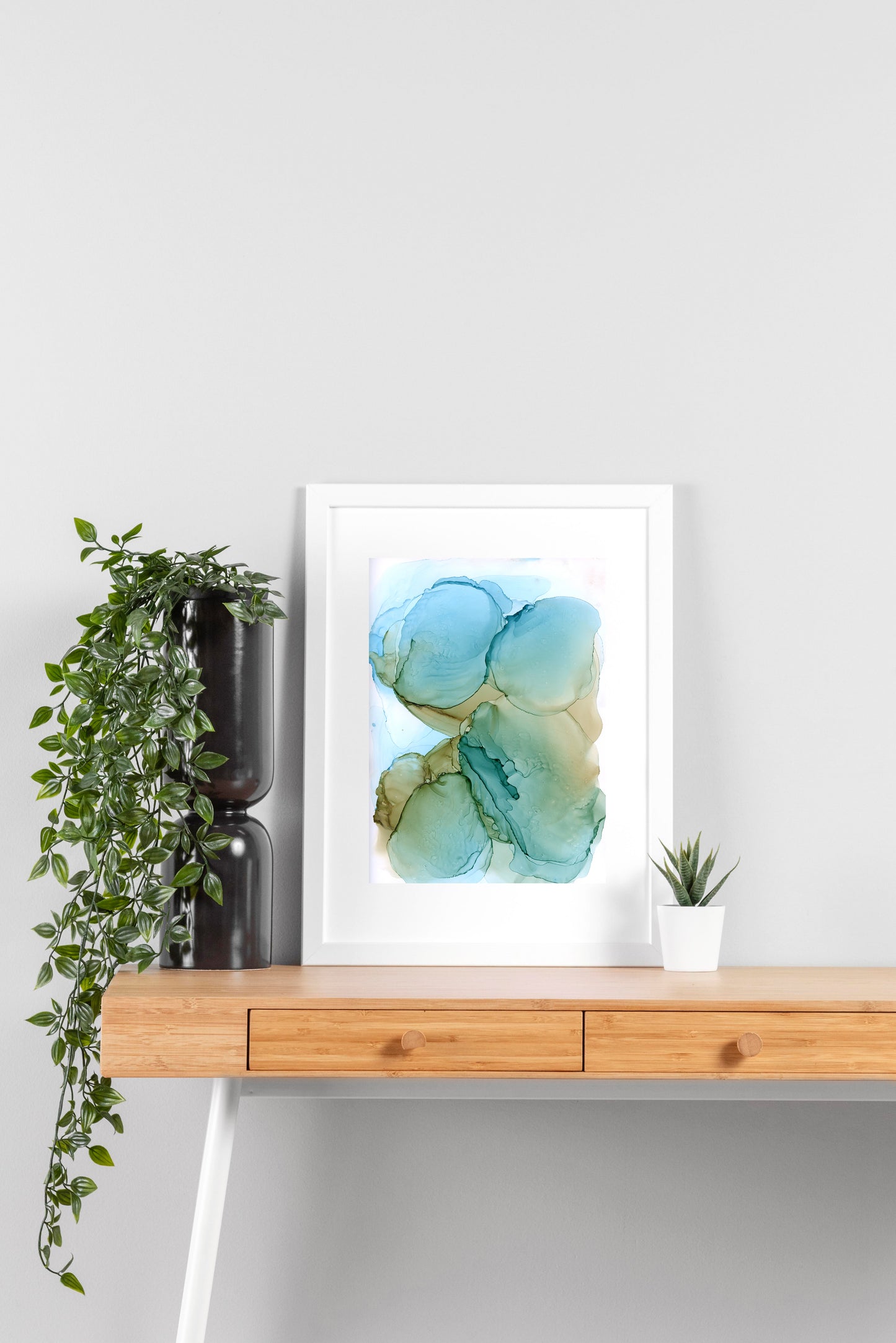 Digital mockup of River Stones framed in white on a wooden desk, leaning up against a neutral wall with a small succulent in a white planter on one side and greenery in a taller black planter on the other.