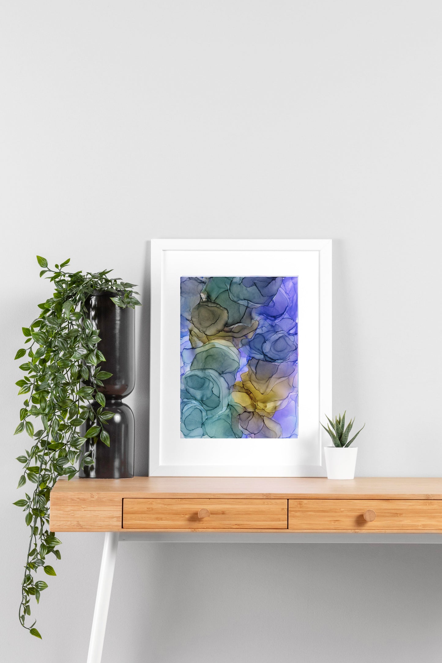 Digital mockup of Moonglade framed in white on a wooden desk, leaning up against a neutral wall with a small succulent in a white planter on one side and greenery in a taller black planter on the other.