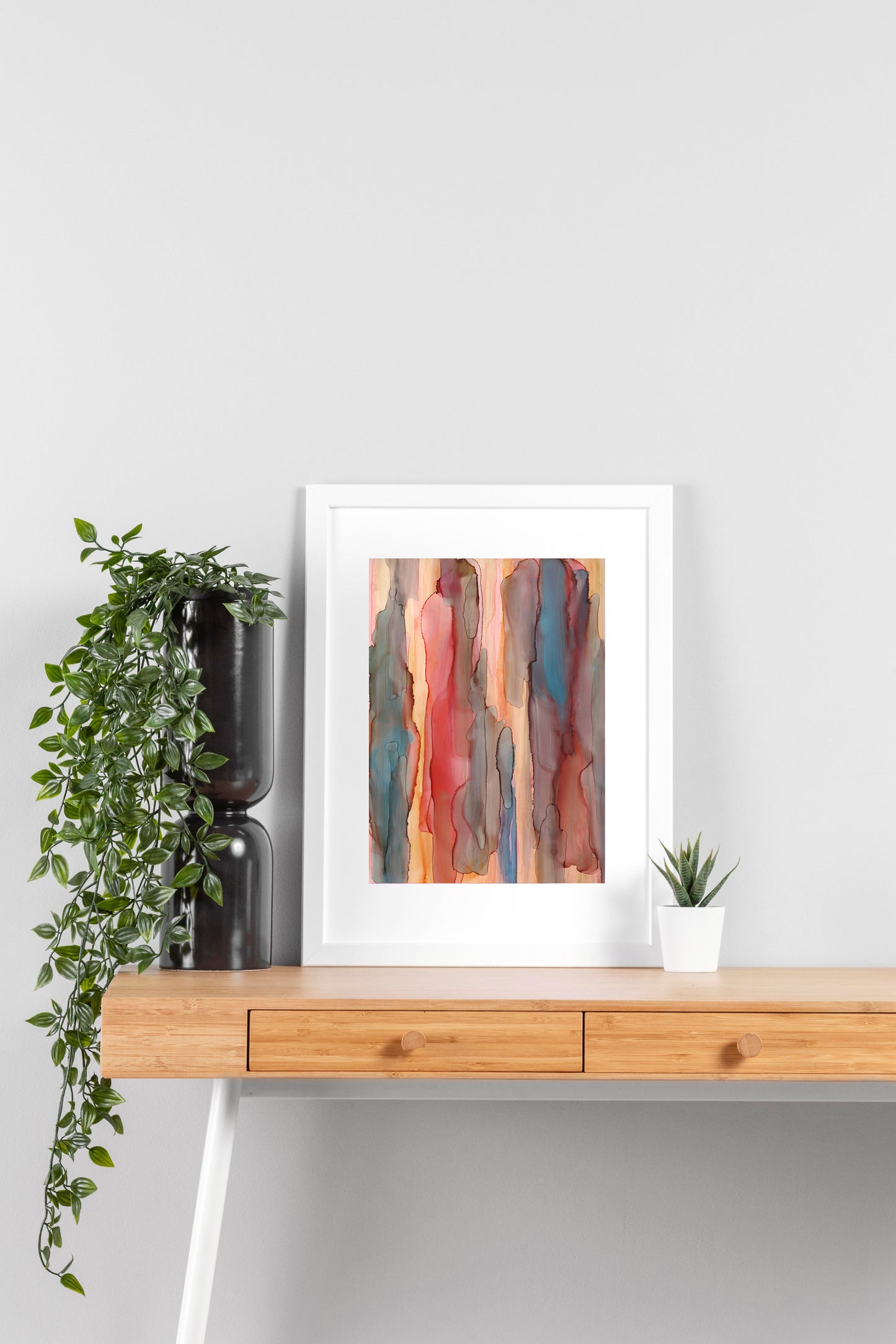 Digital mockup of Playful Reeds framed in white on a wooden desk, leaning up against a neutral wall with a small succulent in a white planter on one side and greenery in a taller black planter on the other.