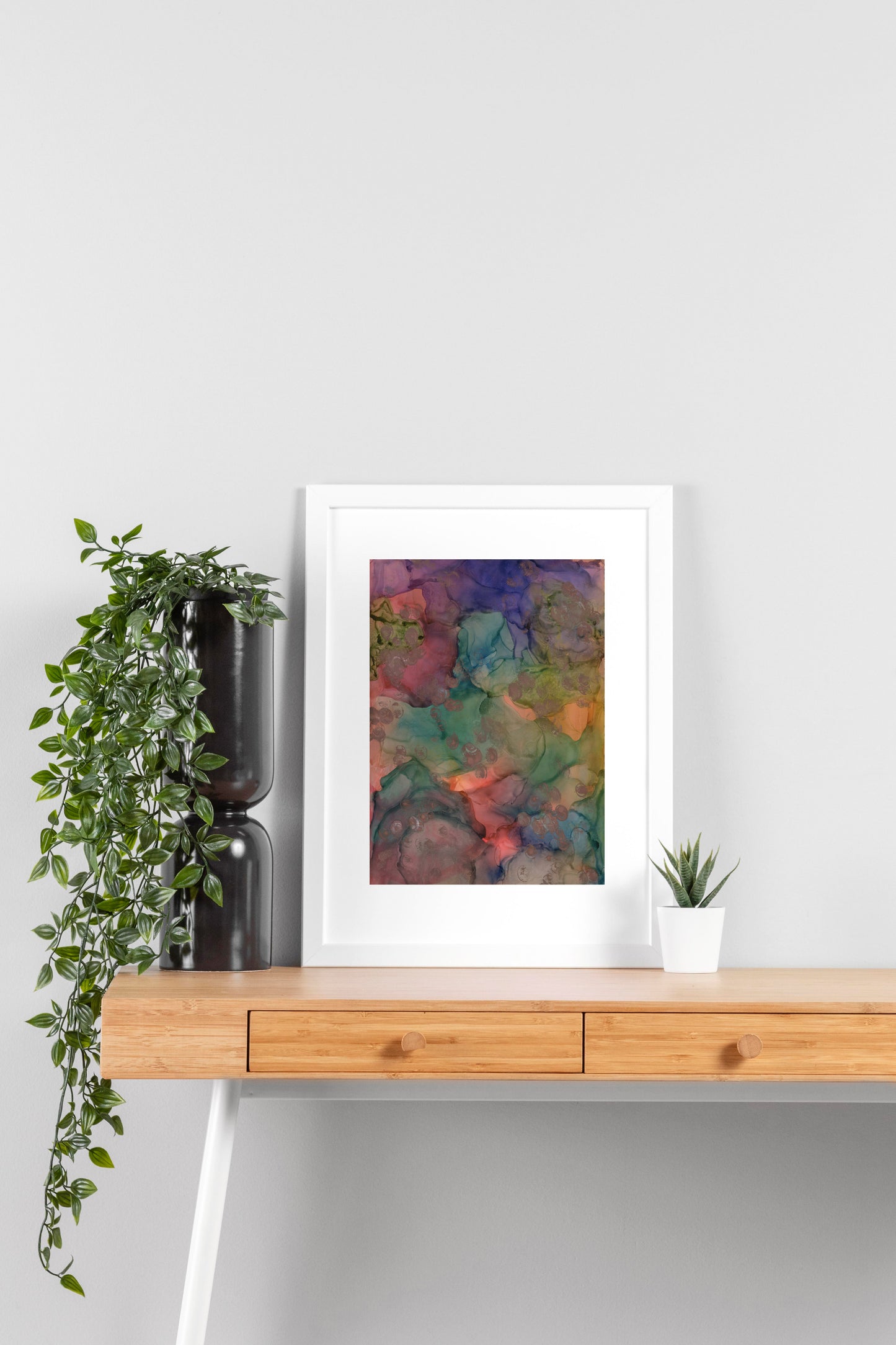 Digital mockup of Coming to the Surface framed in white on a wooden desk, leaning up against a neutral wall with a small succulent in a white planter on one side and greenery in a taller black planter on the other.