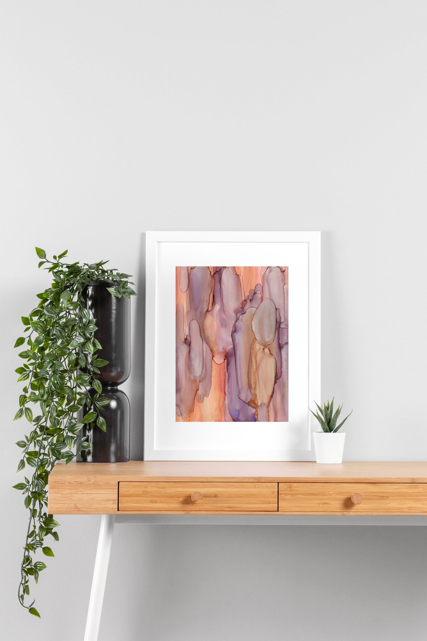 Digital mockup of Sunlit Bay framed in white on a wooden desk, leaning up against a neutral wall with a small succulent in a white planter on one side and greenery in a taller black planter on the other.