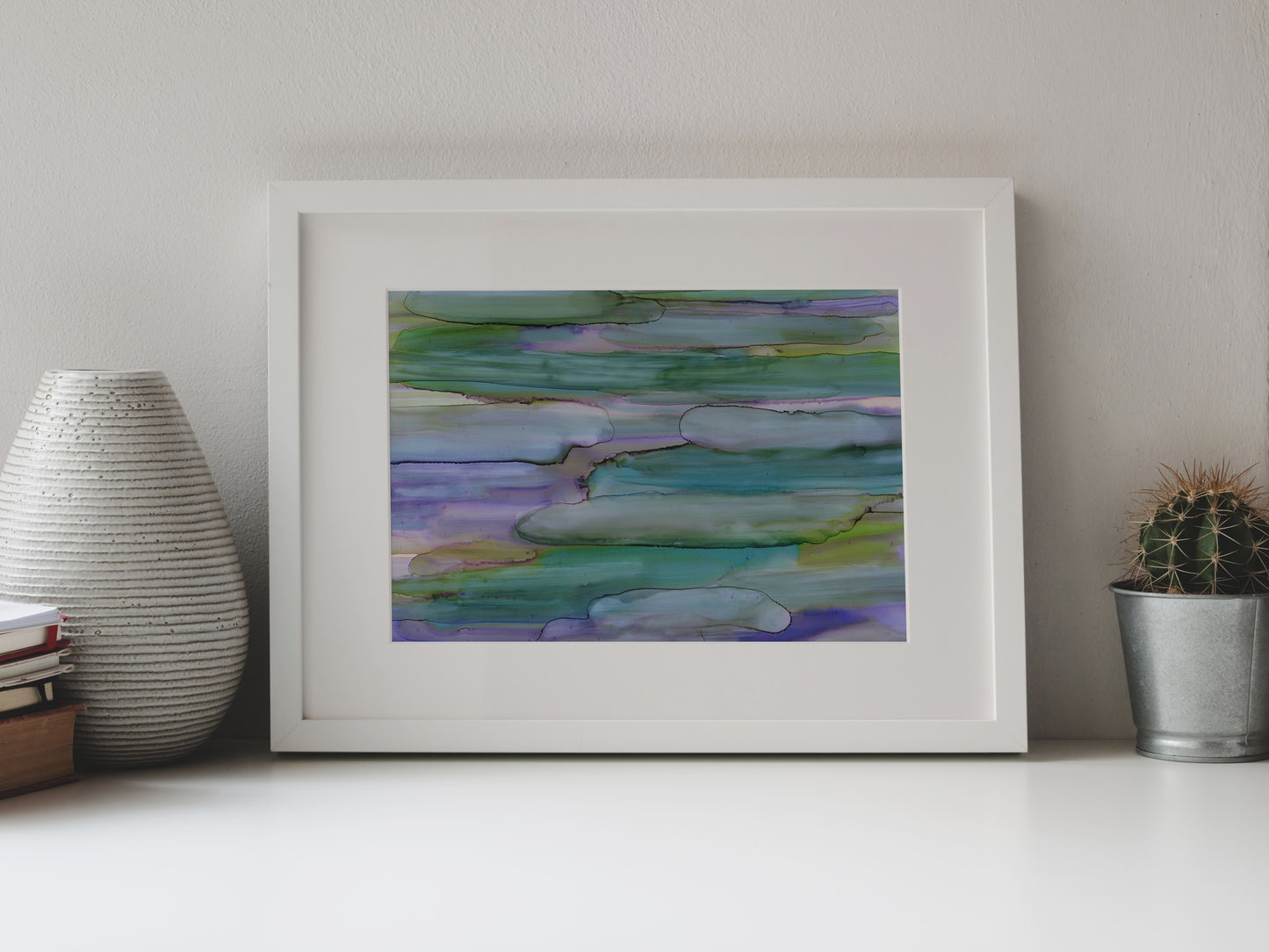 Digital mockup of Interconnected Meadow framed in white on a white table top, leaning up against a white background, surrounded by a vase, stack of books, and tiny cactus in a pail.