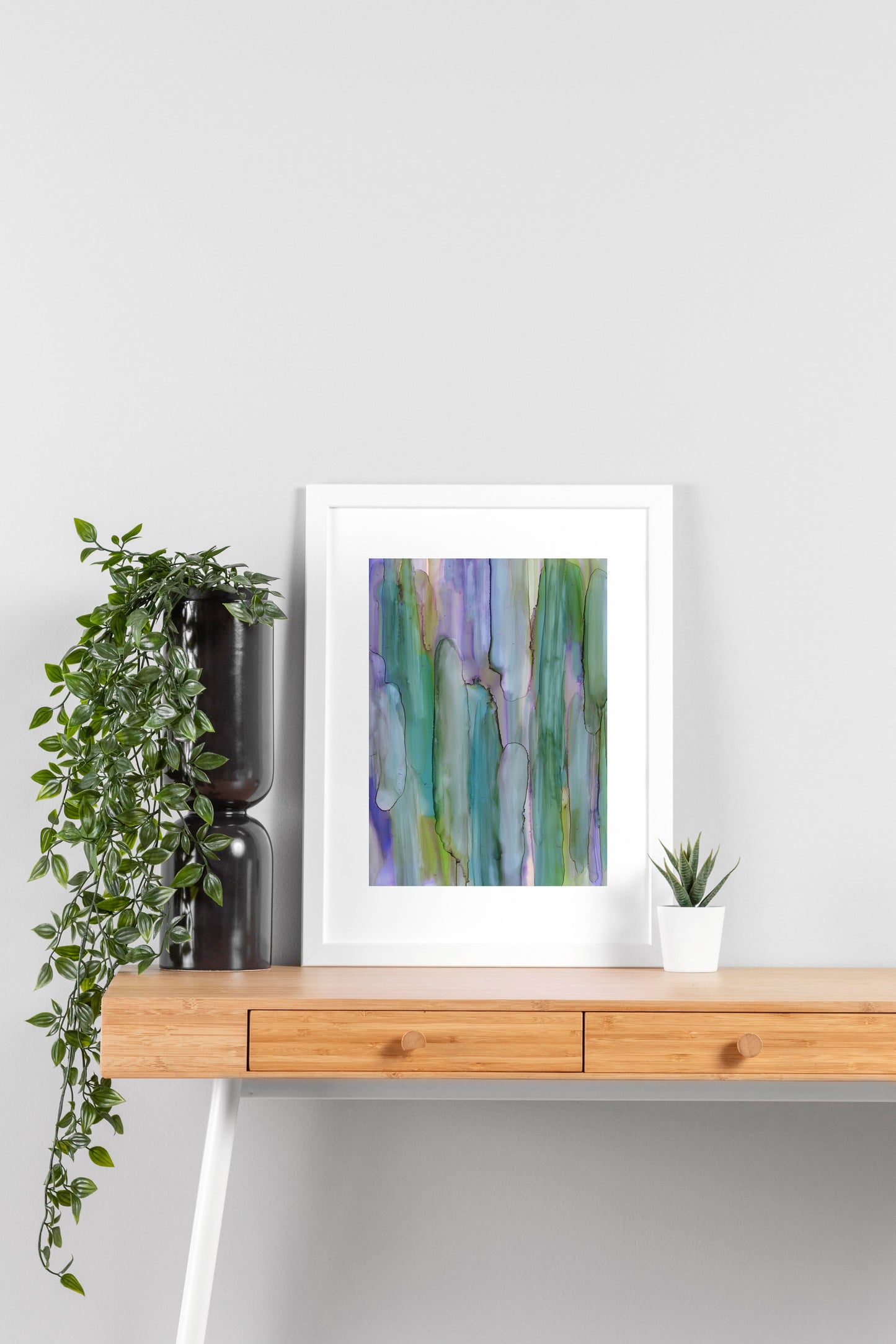 Digital mockup of Interconnected Meadow framed in white on a wooden desk, leaning up against a neutral wall with a small succulent in a white planter on one side and greenery in a taller black planter on the other.