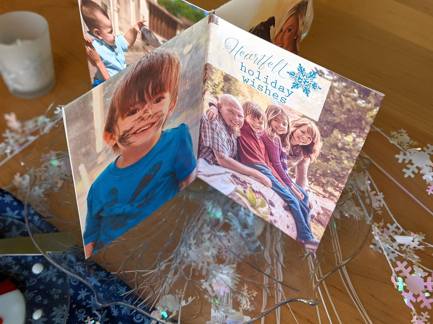 Centerpiece sitting on a glass dessert tray among a snowman plate, snowflake fabric, and snowflake confetti. The panels show a child with a messy face, and a family of four portrait with the words Heartfelt Holiday Wishes.