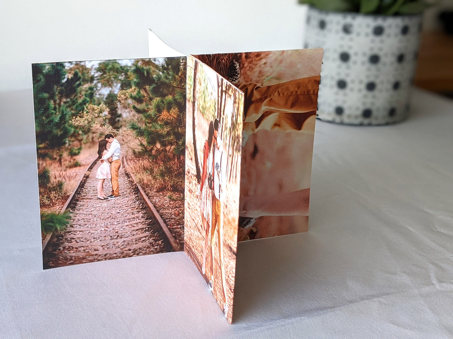 Centerpiece sitting on a white table cloth with a patterned planter in the background. The panels have engagment photos of a couple along an abandoned railroad track and in a wooden area.