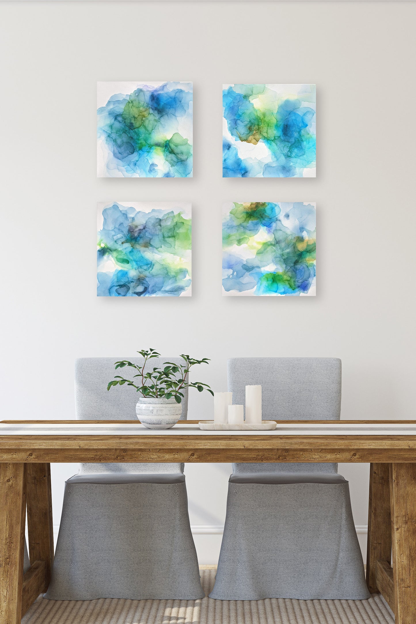 Digital mockup of four canvas hanging on a white modern dining room wall with farmhouse style wooden table and two fabric covered chairs in the foreground. A ceramic planter with green houseplant and tray with three white pillar candles it on the white fabric table runner.