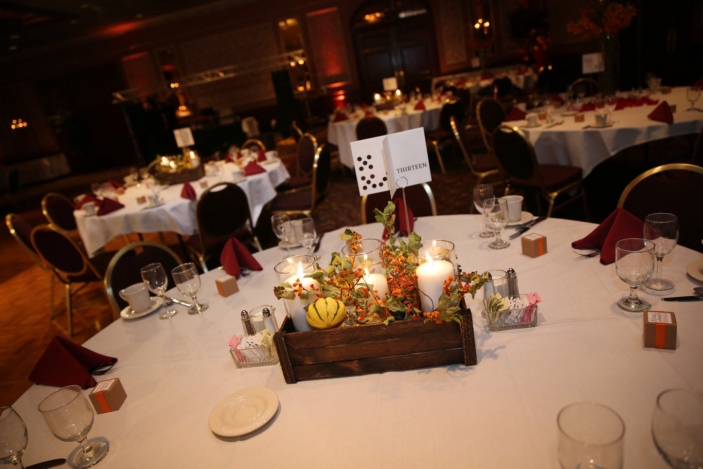 A centerpiece created as a table designation with different representations of the number like domino dots and the written word. It sits above the banquet table with a pick tucked into a wooden box that has fall plants and gourds along with three lit candle votives.