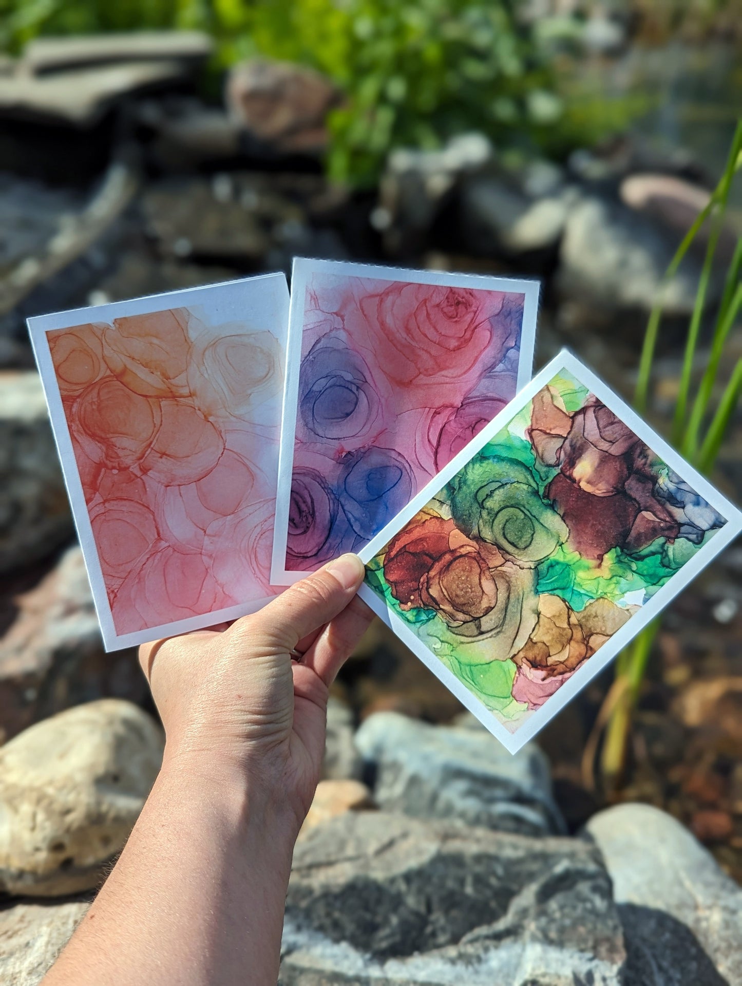 Hand holding 3 greeting cards fanned out with a back drop of rocks and pond waterfall.