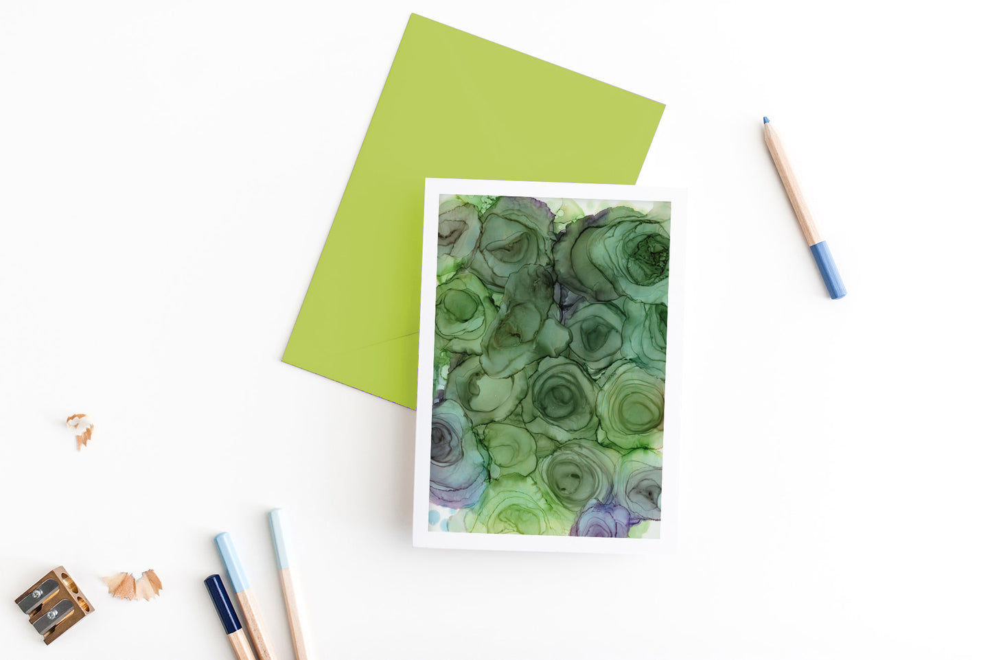 Greeting card, seen from above, with a colored, square flap envelope, pencils, pencil sharpener and shavings on a white background.