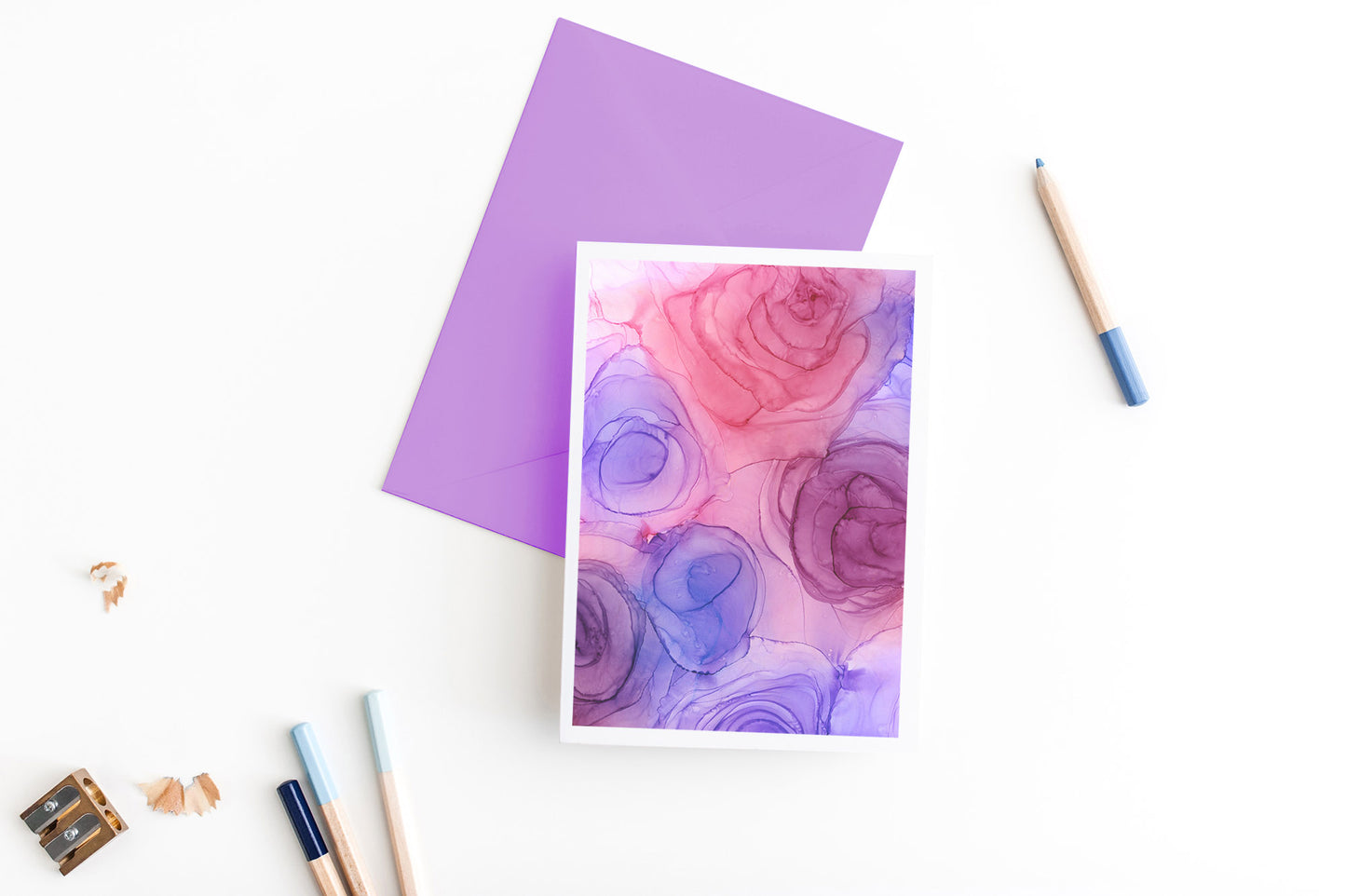 Greeting card, seen from above, with a colored, square flap envelope, pencils, pencil sharpener and shavings on a white background.
