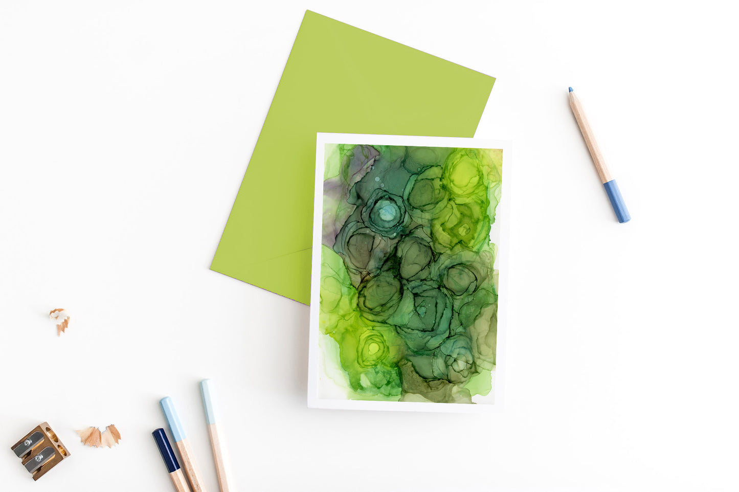 Greeting card, seen from above, with a colored, square flap envelope, pencils, pencil sharpener and shavings on a white background.