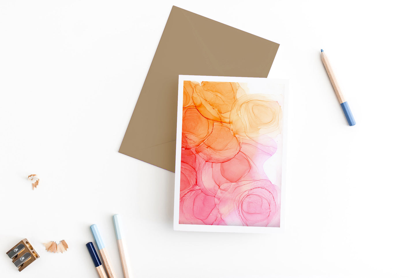 Greeting card, seen from above, with a colored, square flap envelope, pencils, pencil sharpener and shavings on a white background.