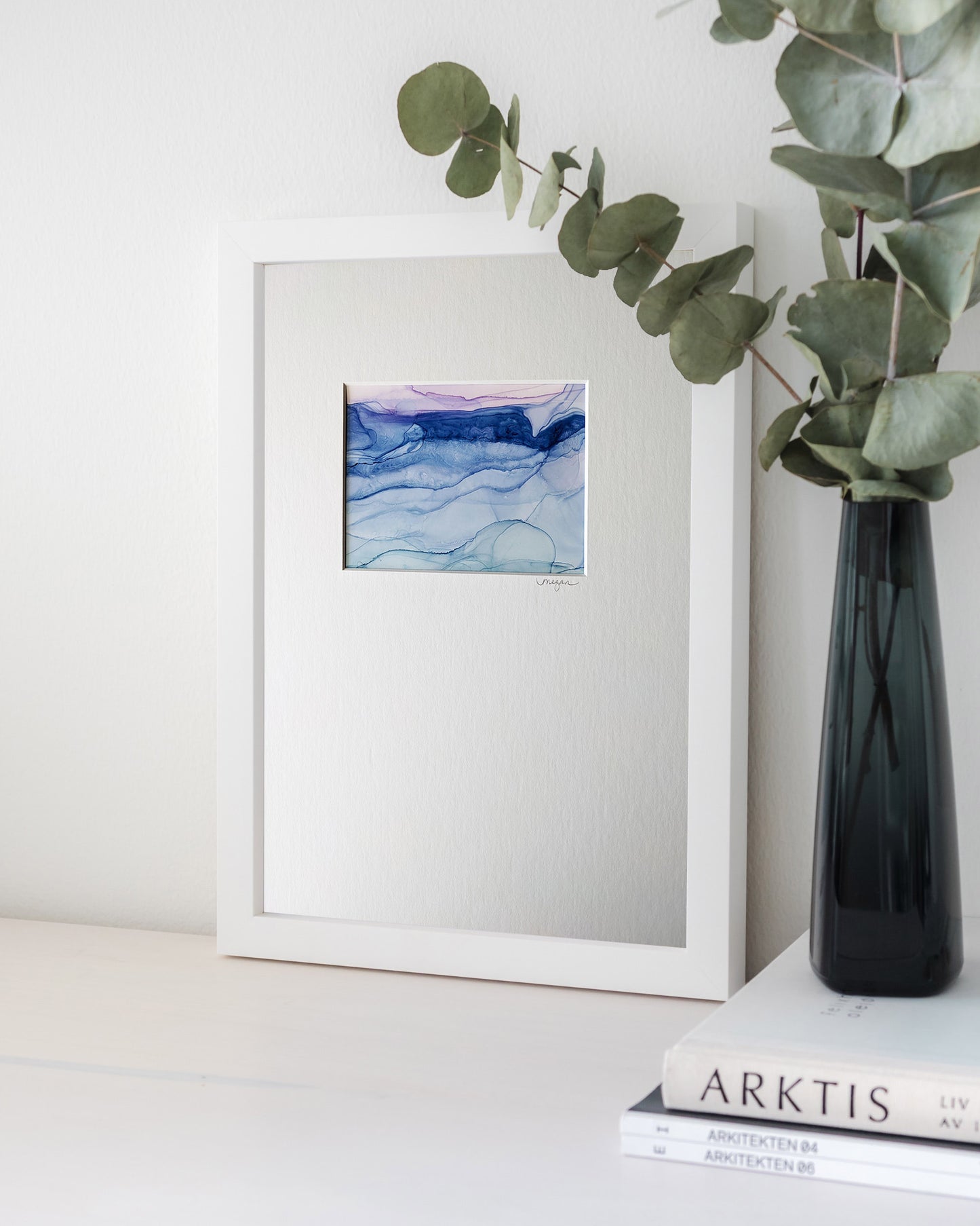 Digital mockup of the painting framed in white leaning against a white background. A black glass vase stacked on magazines holds some eucalyptus leaves off to the side.
