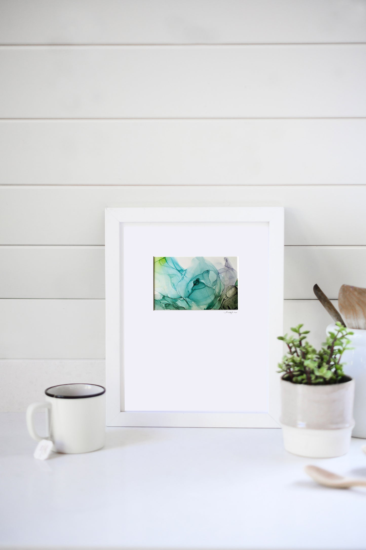 Digital mockup of the painting framed in white leaning against a horizontal white beadboard wall. A ceramic mug, ceramic planter with greenery, and wooden utensils sit to the sides.