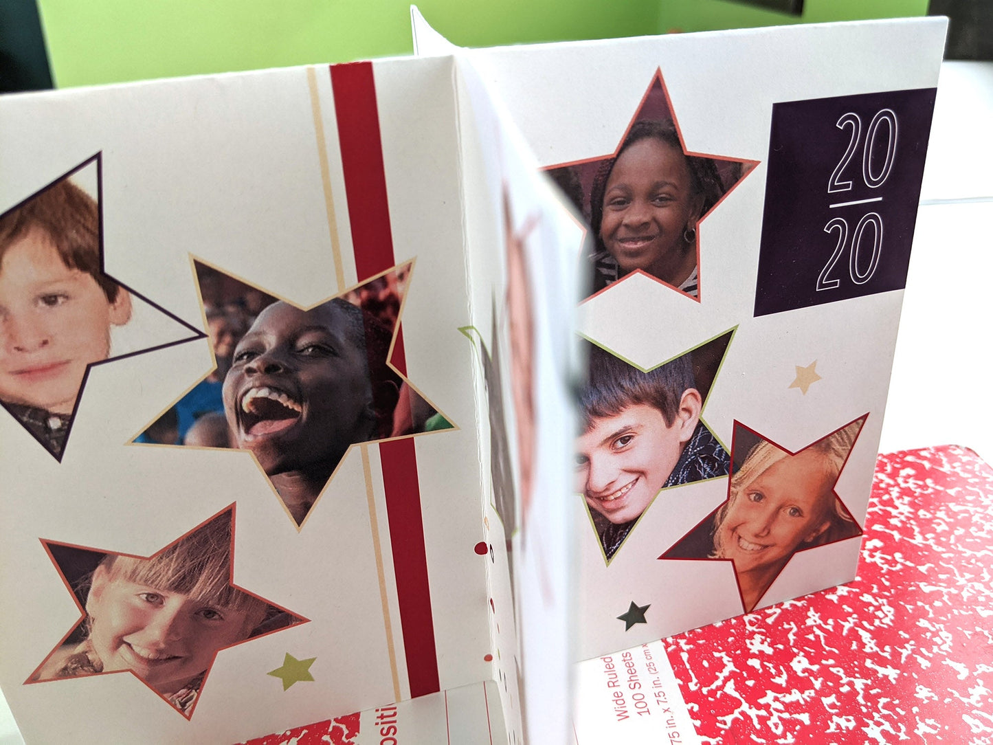 Centerpiece sitting on a red composition notebook with a felt tip pen and small erasers. The panels show photos of children inside colorful stars and the year graphically arranged as a personal keepsake thank you.
