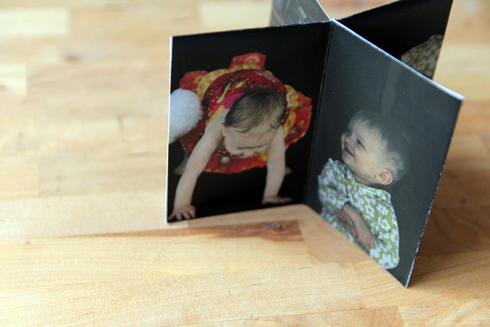 Overhead view of a centerpiece standing on a wooden table top. The photos show a toddler sitting smiling at someone off camera and pushing up to a stand.