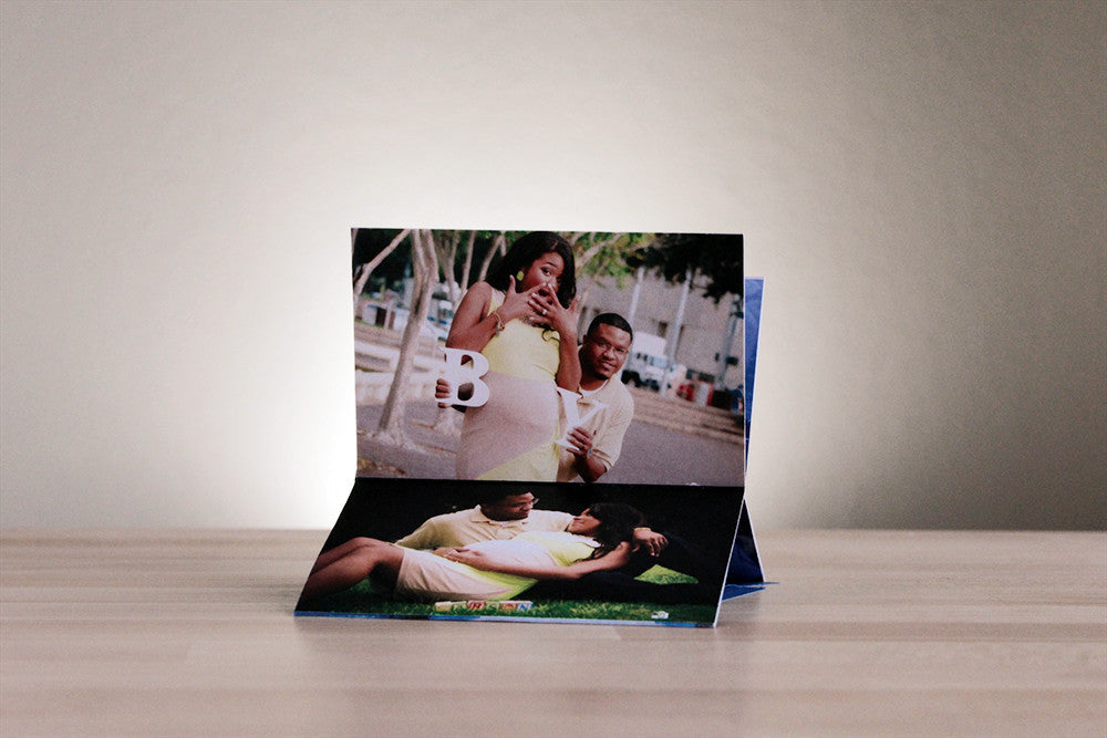 Centerpiece standing on end on a wooden table where the panels show a pregnant person and their partner acting surprised using the belly as the O in BOY as well as them reclining on the grass smiling at each other.
