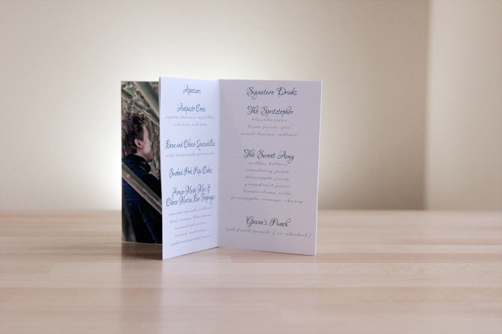 Centerpiece standing on a wooden table with a white wall in the background. The panels show part of an engagement photo, wording for appetizers, dinner menu and specialty drink choices.
