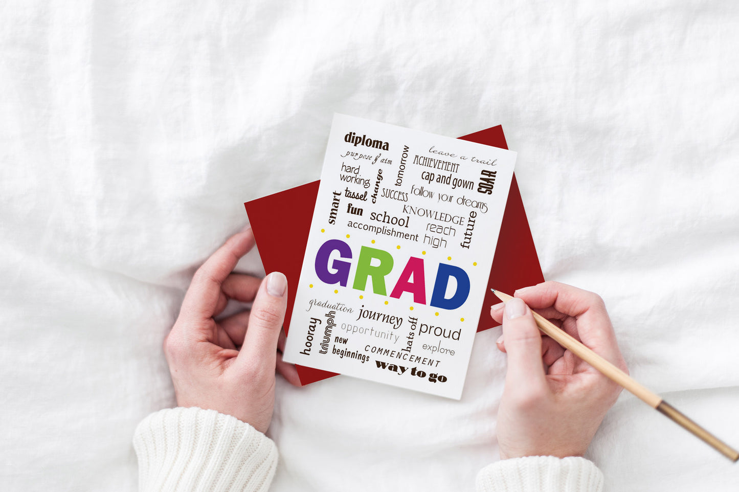Greeting card and colorful envelope, seen from above, held by two hands along with a pencil, ready to write, against a white comforter.