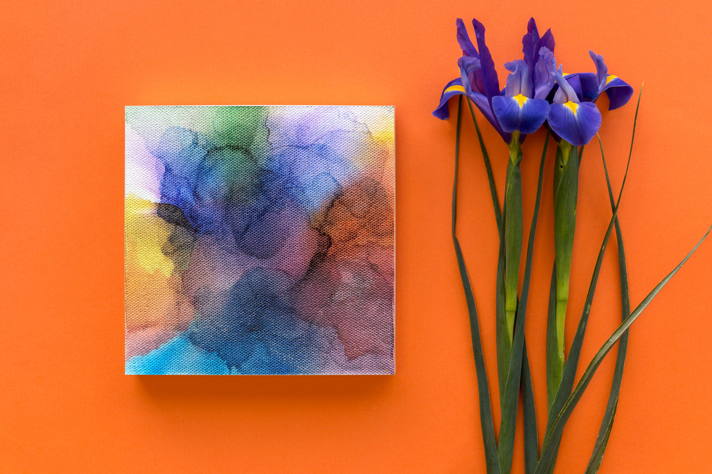 Digital mockup of the canvas laying on an orange backgorund next to stems of purple flowers.