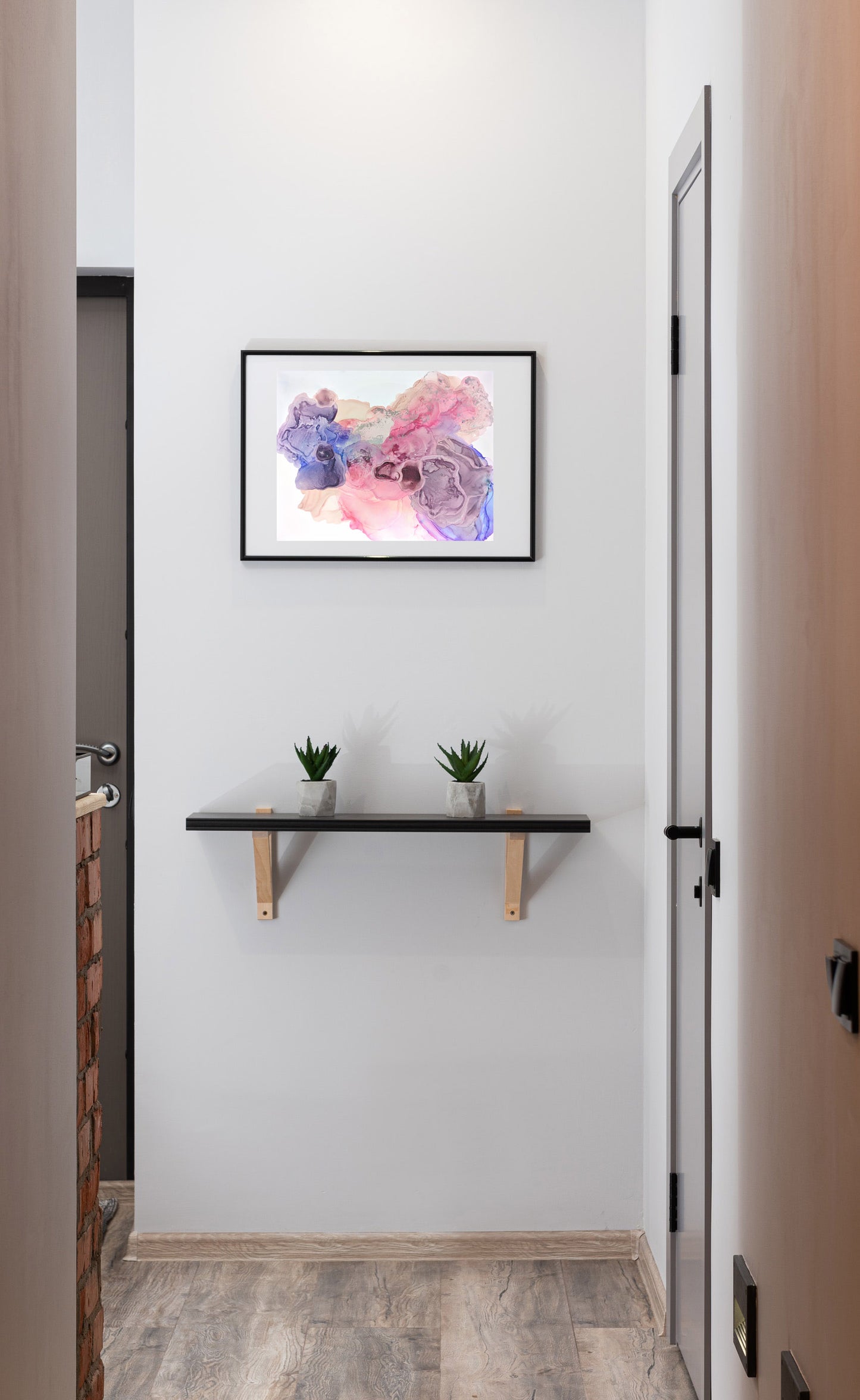 Digital mockup of Available for Joy framed in black hanging on a white wall at the end of a hallway. A wooden shelf with two small succulents in concrete planters sit below.