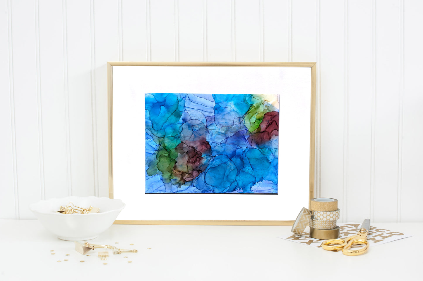 Digital mockup of Balancing Ease and Effort framed with a thin gold frame standing on a white counter leaning against a white beadboard wall. There are gold and white accessories - washi tape, scissors, stickers binder clips and confetti on either side.