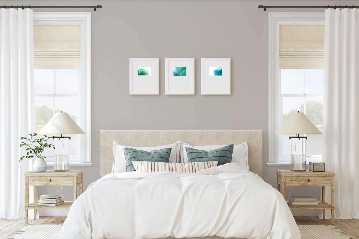 Digital mockup of three paintings framed in white against a light grey wall in a bedroom between two windows with a bed between two end tables in the foreground.