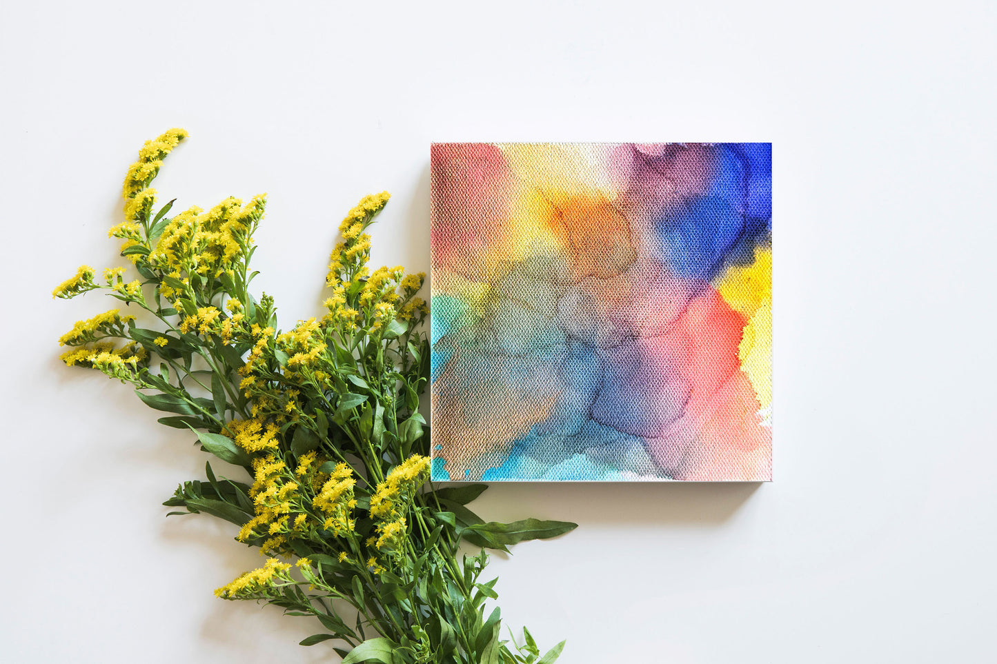 Digital mockup of the canvas laying on a white backgorund next to stems of tiny yellow flowers.