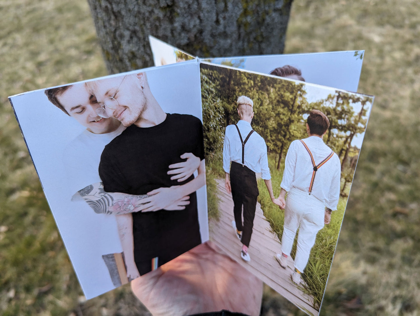 Hand holding a centerpiece with an LGBTQ+ couple with arms wrapped around each other and heads tilted together and another photo from behind with them walking hand and hand down a boardwalk in a forest.