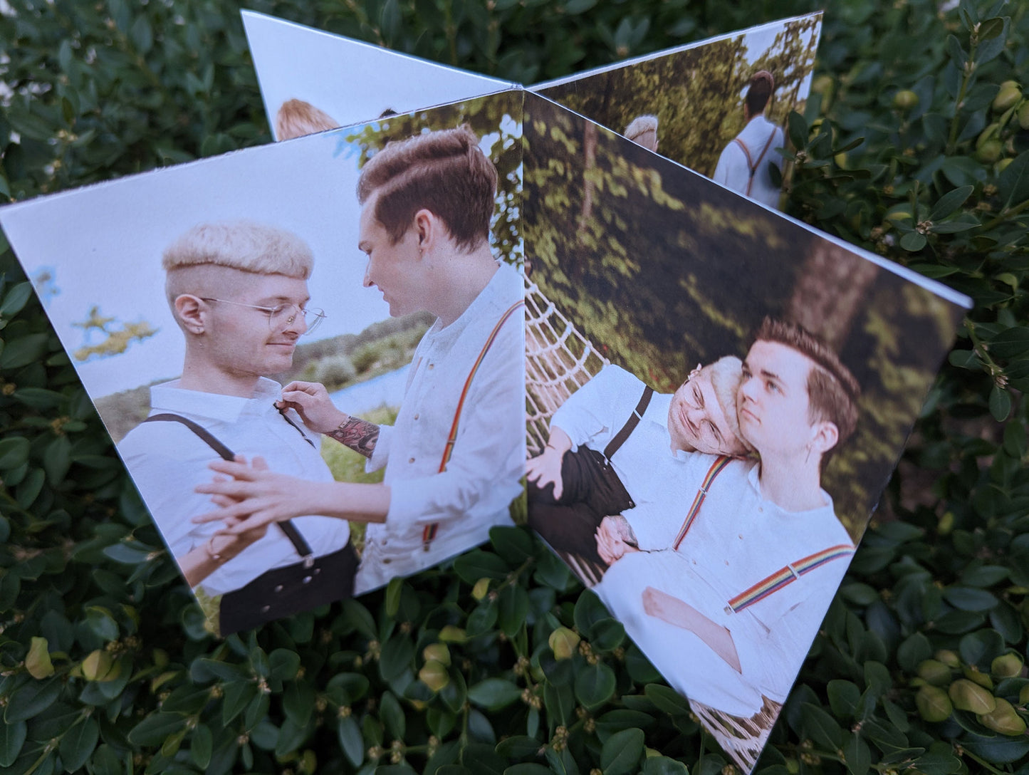 Centerpiece seen from above sitting on a green shrub showing photos of an LGBTQ+ couple in white shirts and suspenders facing each other and holding hands and sitting next to each other in a hammock heads resting together.