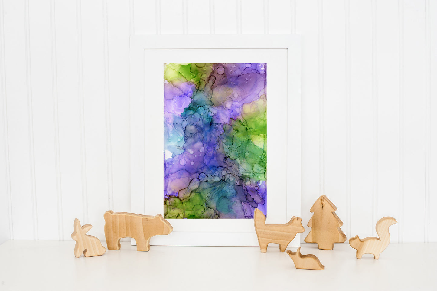 Digital mockup of Claiming Happiness framed in white standing on a white countertop and leaning against a white beadboard wall. There are wooden animal and tree cutouts standing at the base of the piece.