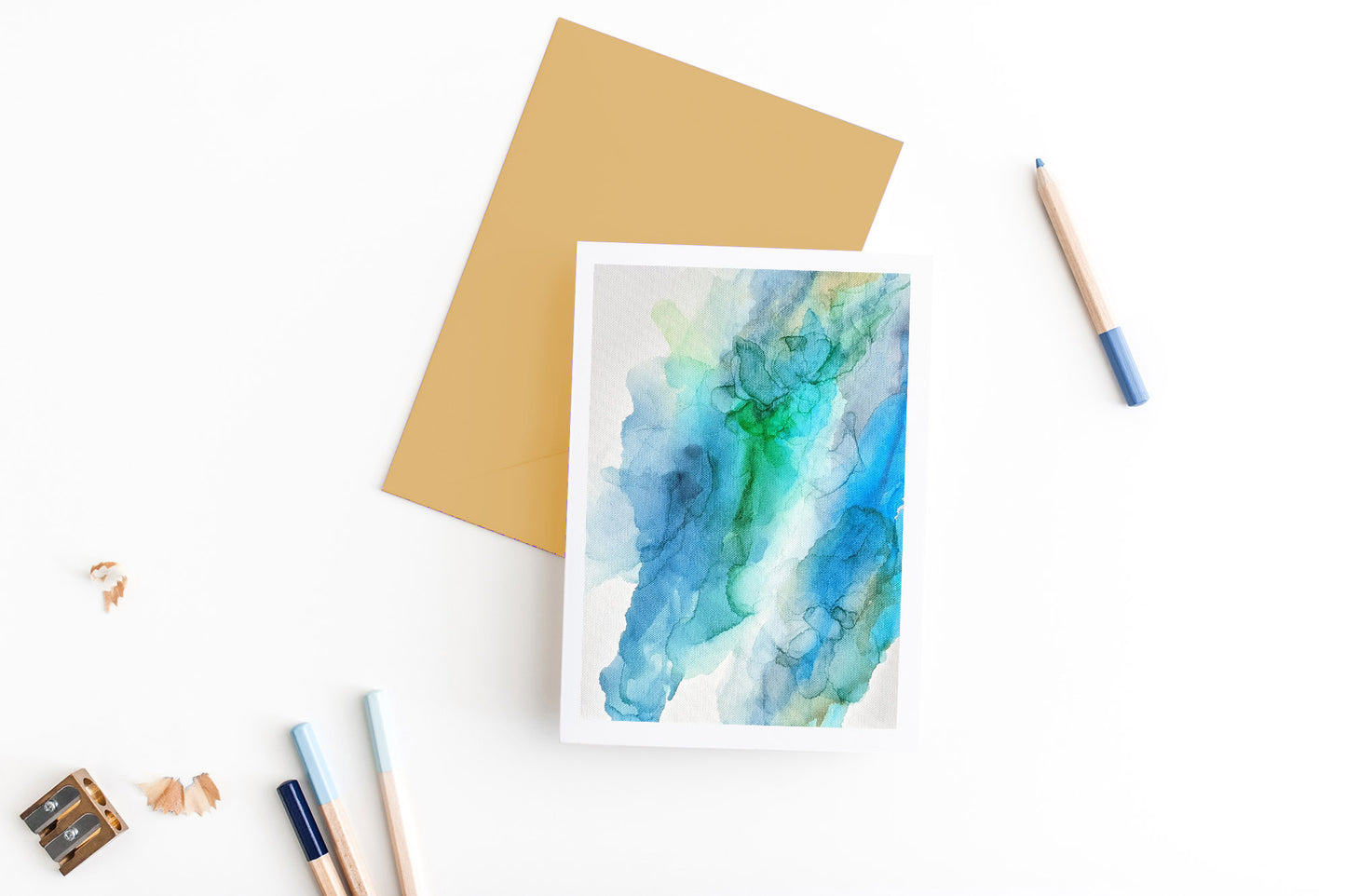 Greeting card, seen from above, with a colored, square flap envelope, pencils, pencil sharpener and shavings on a white background.