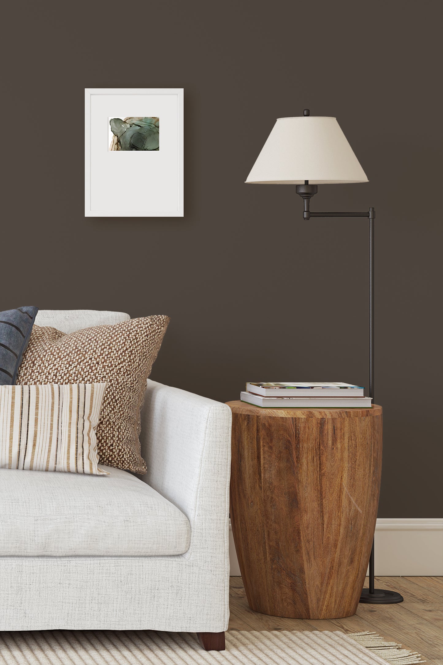 Digital mockup of painting framed in white against a brown wall above the end of a sofa with a modern wooden end table and floor lamp.