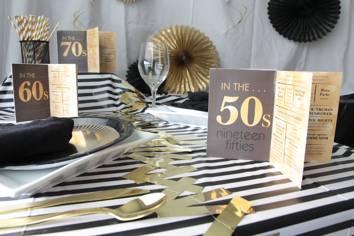 Black and white striped table cloth with square white dinner plates and gold and black party accessories along with black and gold table centepieces celebrating the 50s, 60s, and 70s decades.