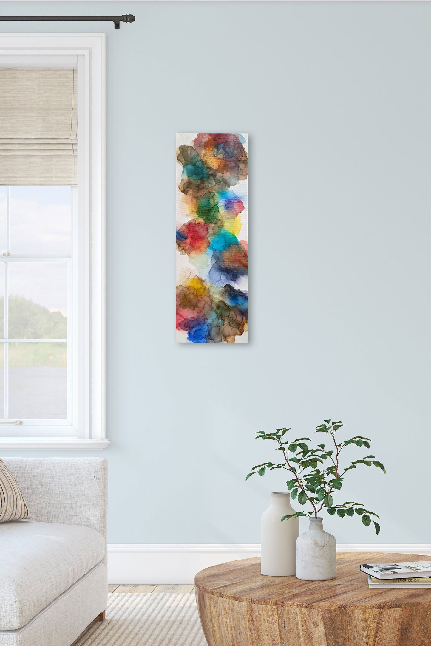 Digital mockup of Expanding Boundaries hung on a light blue living room wall next to a window and edge of sofa. A wooden coffee table with ceramic vases, greenery and a stack of books sits in the foreground.