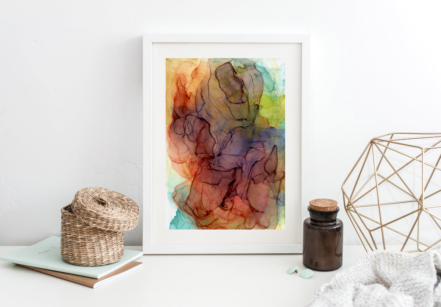 Finding Flow in the Unfolding framed in white leaning against a white background. Small notebooks, a woven basket, an apothocary bottle and a gold sculpture sit off to the sides.