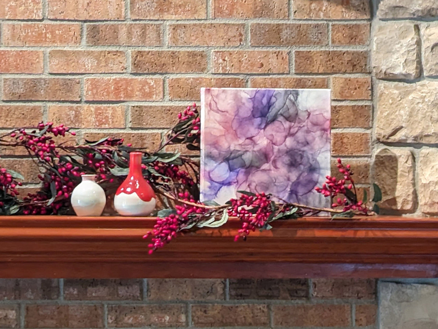 Canvas sitting on a wooden fireplace mantel leaning against a brick wall with ceramic vases and a berry vine around it.