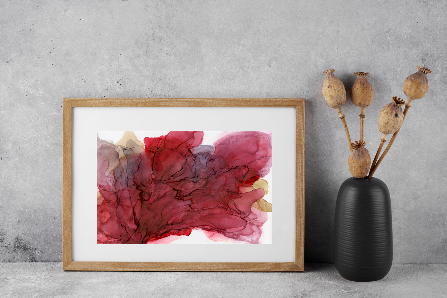 Digital mockup of Inviting Love and Joy framed in wood sitting on a grey surface leaned against a grey concrete background. A black ceramic vase with pods on stems sits to the side.