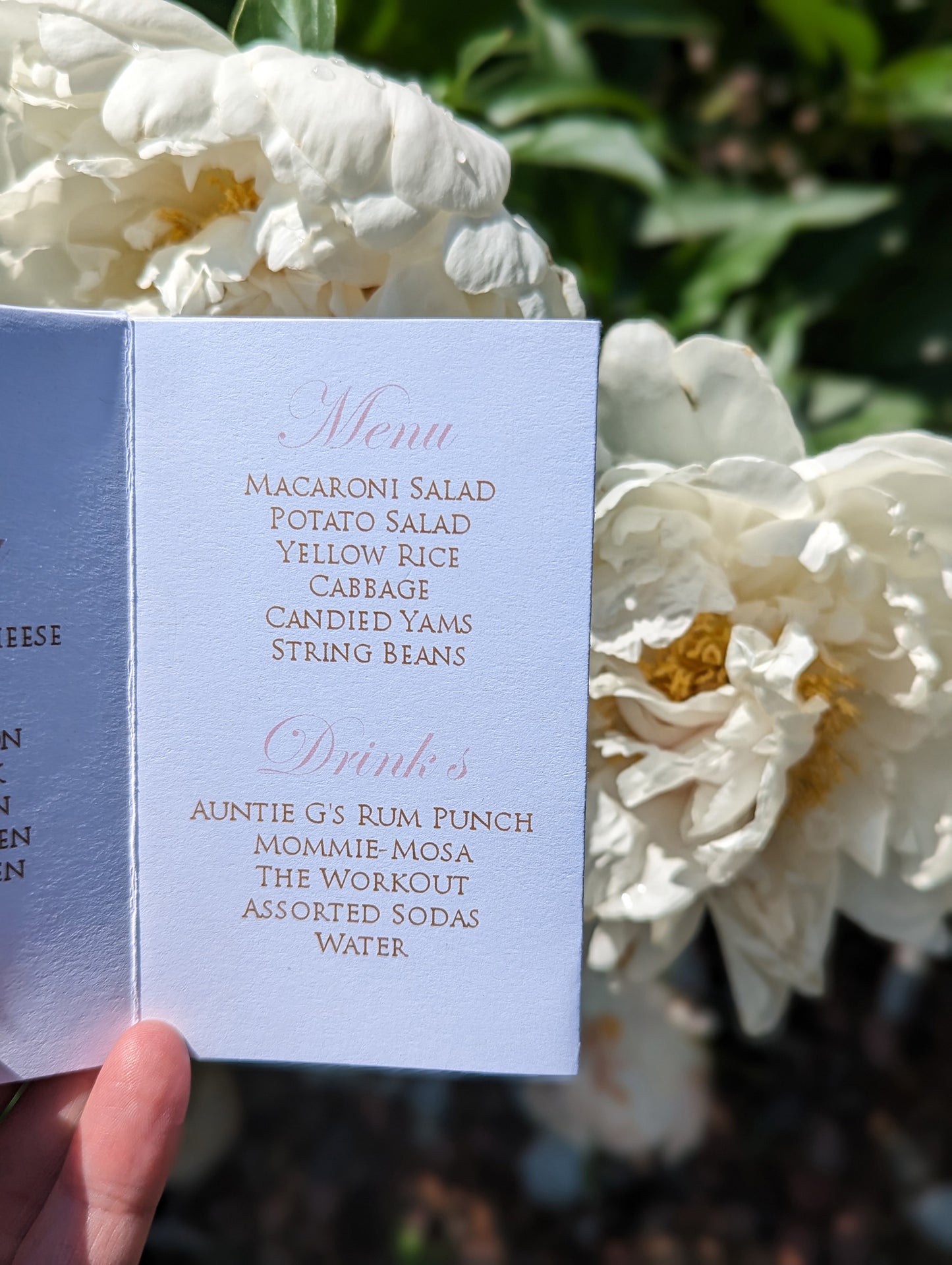 Hand holding a centerpiece showing a panel with a menu and drink choices. Large white peony flowers and greenery fill the background.